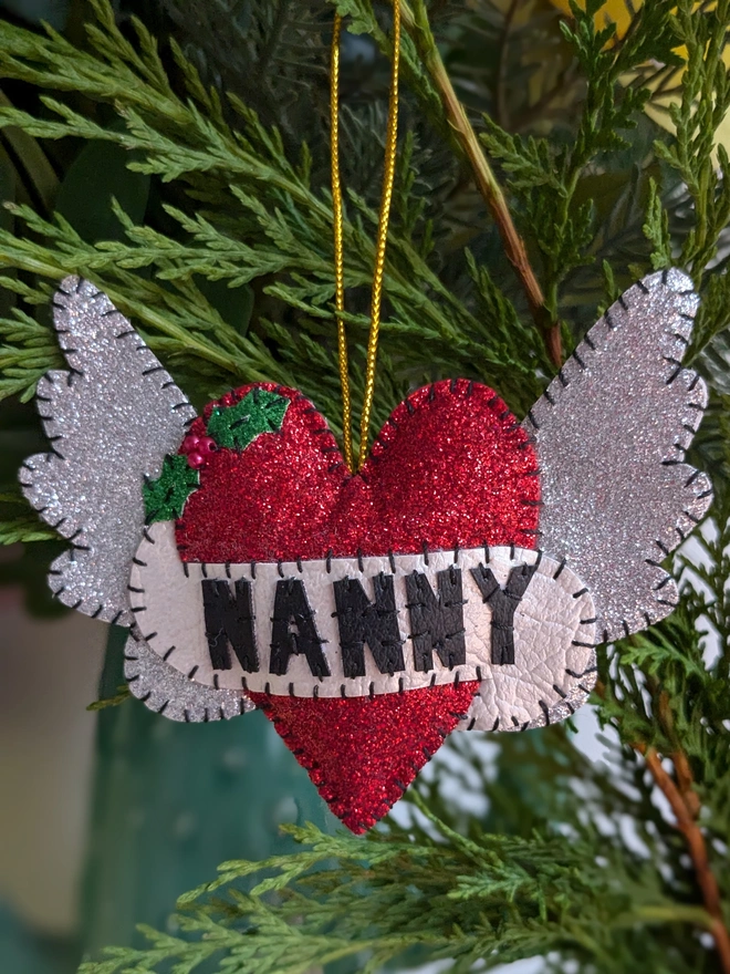 Personalised hand-stitched red glitter heart Christmas decoration with the name ‘NANNY’, silver glitter angel wings, holly detail and a gold hanging loop, displayed amongst festive greenery.