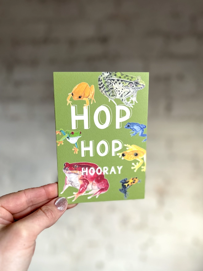 a green greetings card featuring a selection of frog illustrations with the text “hop hop hooray”