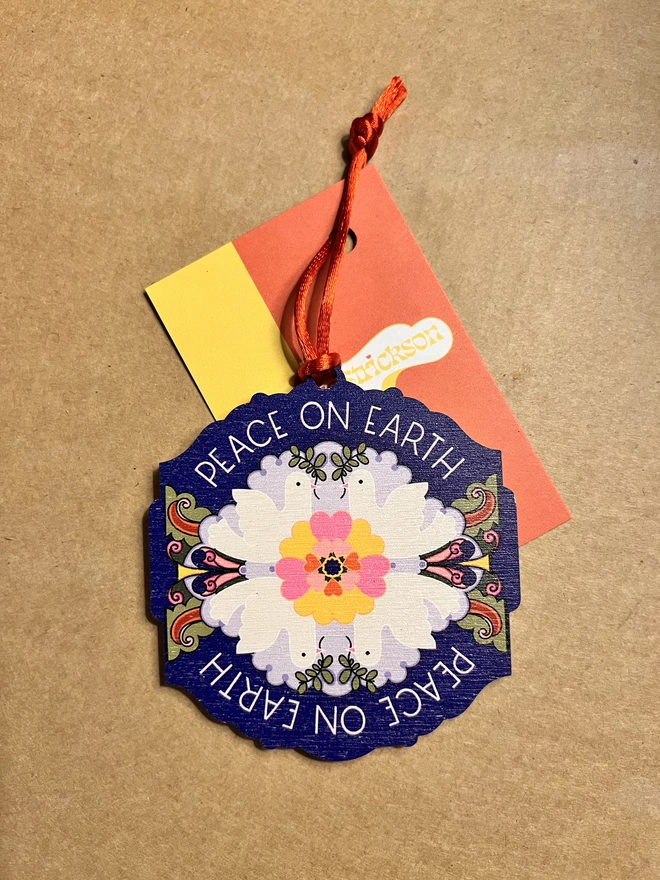 Multi coloured wooden tree decoration the reads 'Peace on Earth' around it, with four white doves and a flower pattern at the centre. It is lying on a brown card background with a red ribbon and small business card that says 'Rebecca Strickson'