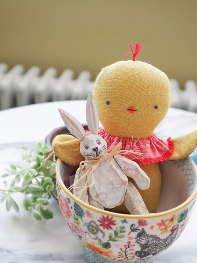 Handmade chick doll made from soft yellow fabric, with a stitched face, small beak, ruffled collar, and patterned fabric arms and long legs, sat in a tea cup with wooden bunny doll.