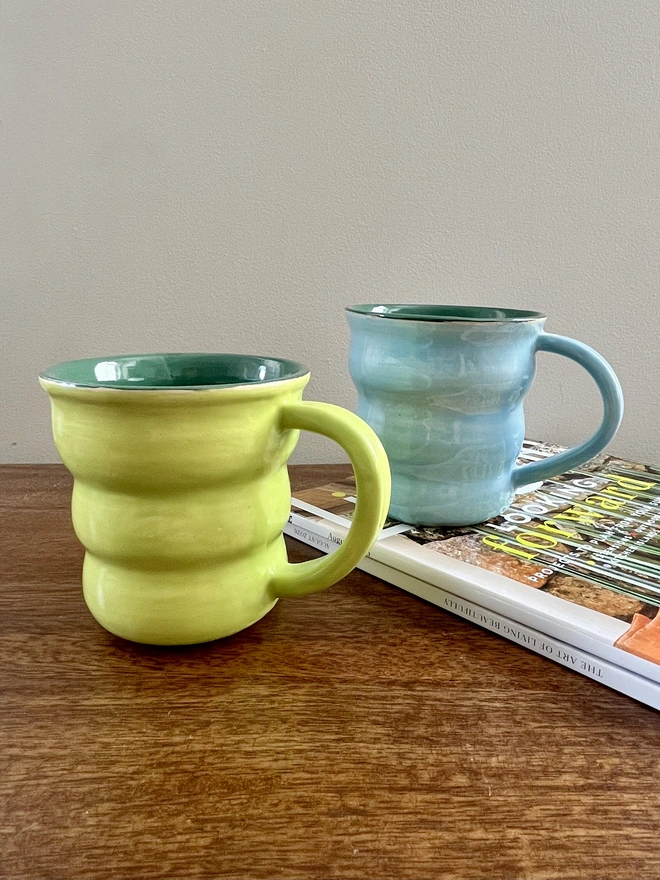 Two hand-thrown 400 ml ceramic mugs with wavy shapes and rounded handles, glazed in lime green and sky blue with forest green interiors, handmade by Hannah Inskip Pottery. Mugs sit on wooden table and stack of magazines