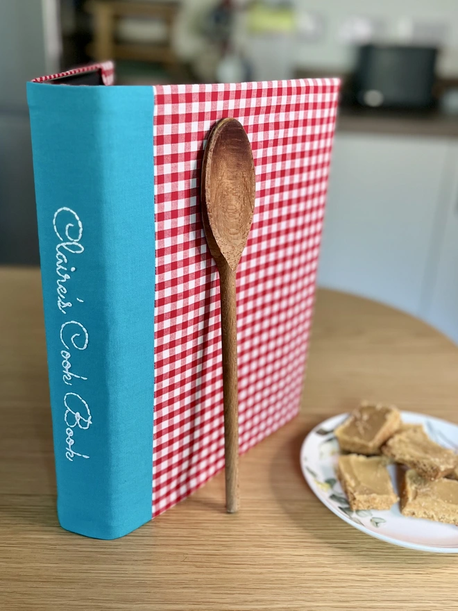 Red gingham recipe binder with 'Claire's Cook Book' embroidered on teal spine, standing on kitchen table with wooden spoon propped against it and a plate of baking beside the folder.