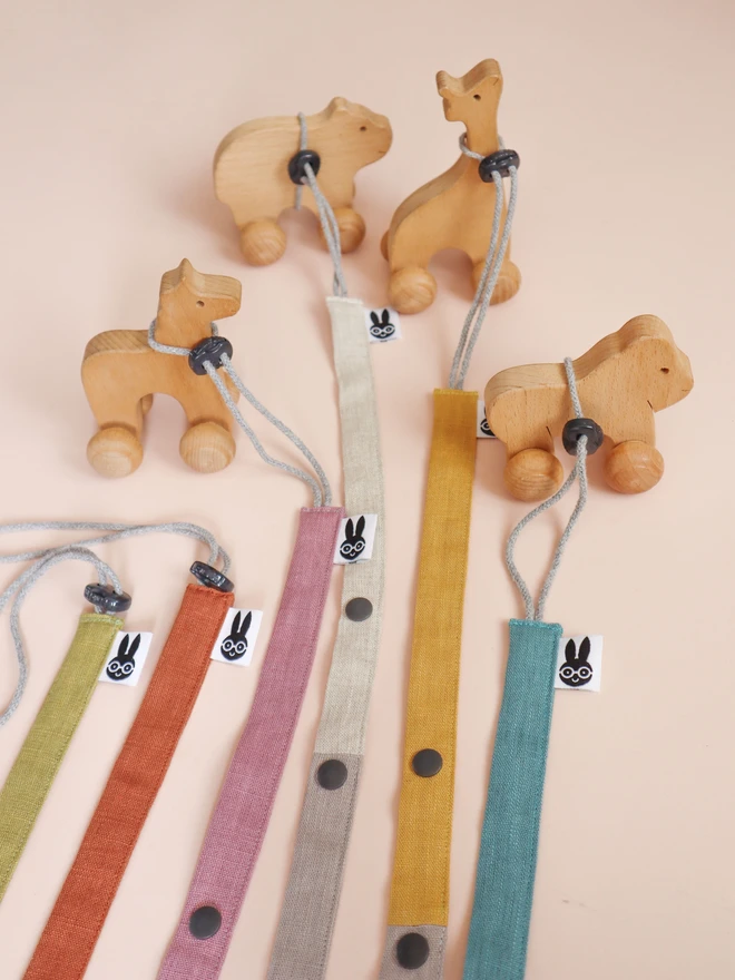 Handmade linen toy strap with grey string and a toggle attached to wooden baby toys, along with other colour options in rainbow colours.