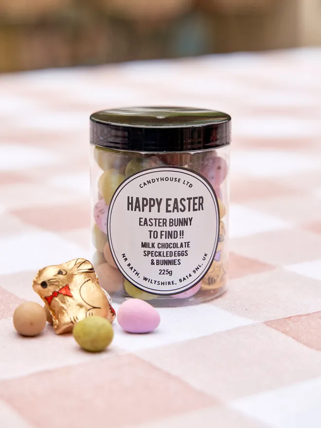 A clear Jar with a black lid filled with speckled eggs and a chocolate easter bunny inside, placed on a pink checkered tablecloth with a few eggs and one bunny by the side of the jar 