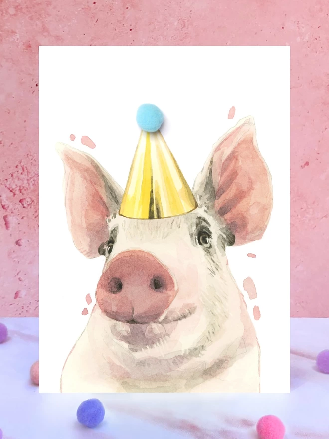 A greeting card featuring a hand painted design of a pig, stood upright on a marble surface surrounded by pompoms. 