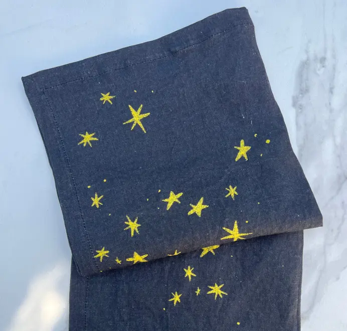 Stars Napkins In White (Set of 4). White cloth napkins with scattered yellow star prints, placed on a Stars Napkins In White (Set of 4). White cloth napkins with scattered yellow star prints, placed on a blue table cloth. 