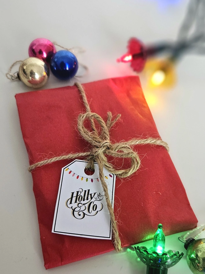 Photograph of the bauble wrapped in red tissue paper with brown twine and a gift tag illustrated with a string of colourful vintage baubles and the Holly and Co logo