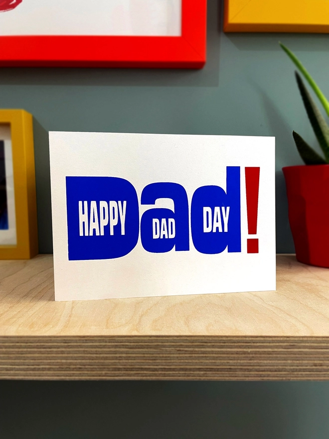 White landscape greetings card on a plywood shelf. The design is typography of the word Dad! With happy Dad Day written within the letters. The writing is printed in royal blue with an oversized exclamation mark in red to the right. Coloured frames can be seen around and a plant to one side.