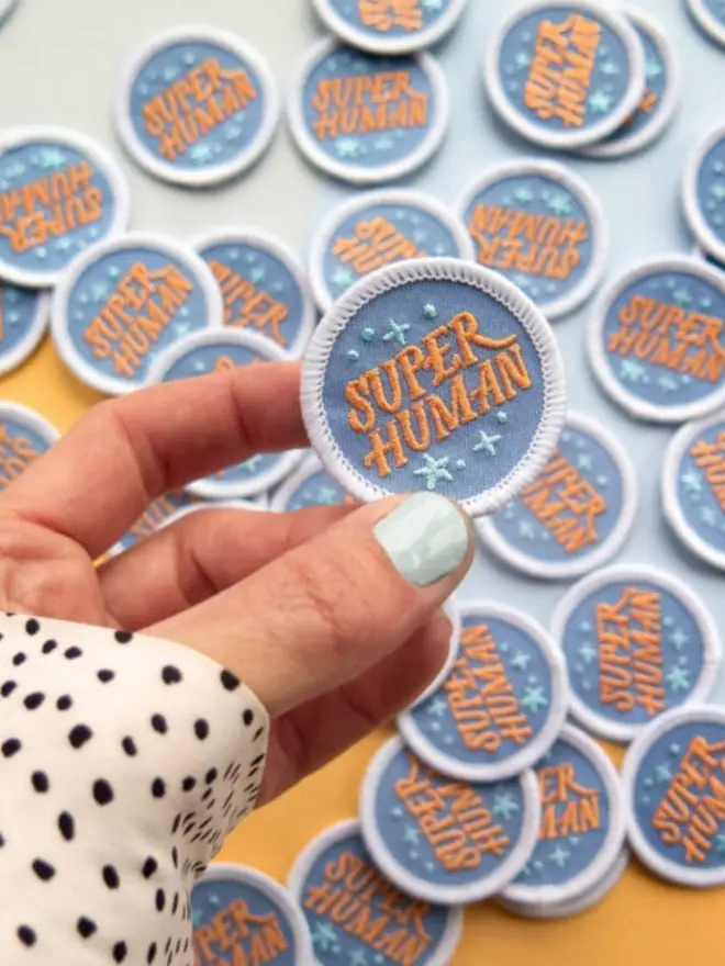 A close-up of a hand holding the round embroidered "Super Human" patch with a white stitched border. More patches are piled up in the background.