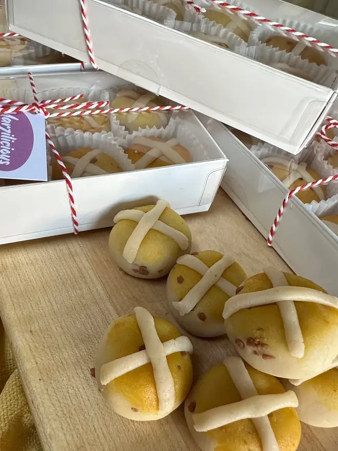 Marzipan Hot Cross Buns (Box Of 6 Or 9), marzipan hot cross buns on a wooden surface in front of more marzipan hot cross buns packaged in the background. 