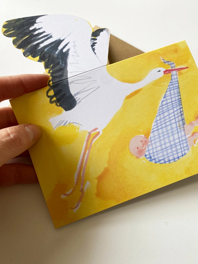A new baby card by Esther Kent showing a black and white stork carrying a baby in a blue checked cloth, on a yellow background