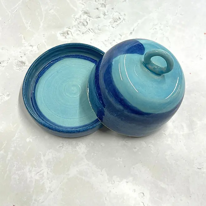 Landscape Butter Dish, a colourful butter dish sitting on a stone surface. In the image the lid is lifting off of the dish. 