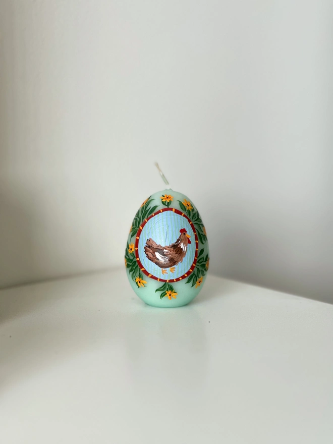 Hand Painted Easter Egg Candle Hen & Flowers, an Easter egg candle sitting on a plain surface. It's Easter and floral design wraps around the egg. 