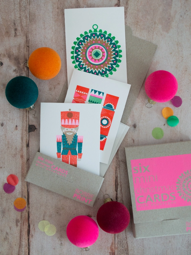 A set of 6 mini Christmas greeting cards lies on a wooden table spilling out of their box while small brightly coloured festive baubles are scattered around. A closed boxed of six mini Christmas cards lies to the side of the open box.