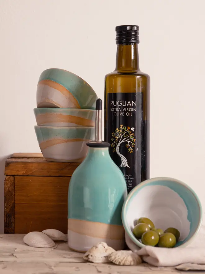 The Shoreline oil pourer surrounded by mini matching bowls, one of which is on its side filled with green olives. Behind the oil pourer is a bottle of olive oil.