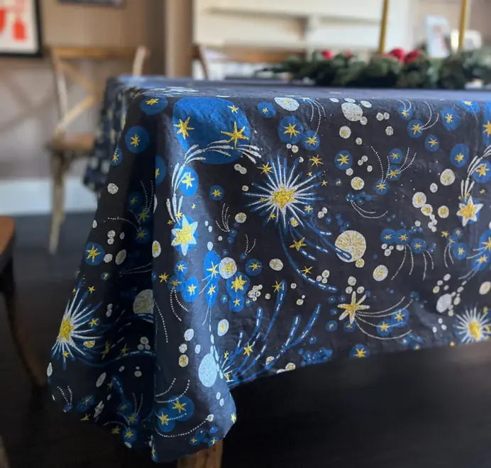 Solaris Tablecloth In White Or Blue. Blue tablecloth with blue and yellow starburst motifs, styled with white plates and a matching napkin.