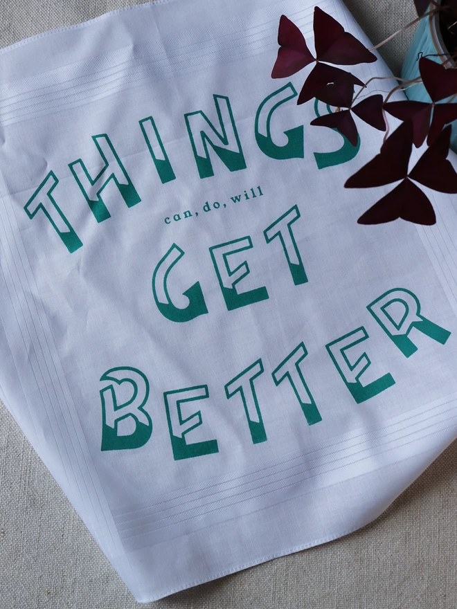 Things Get Better Handkerchief printed in green, laid on a linen tablecloth with a red-leafed houseplant