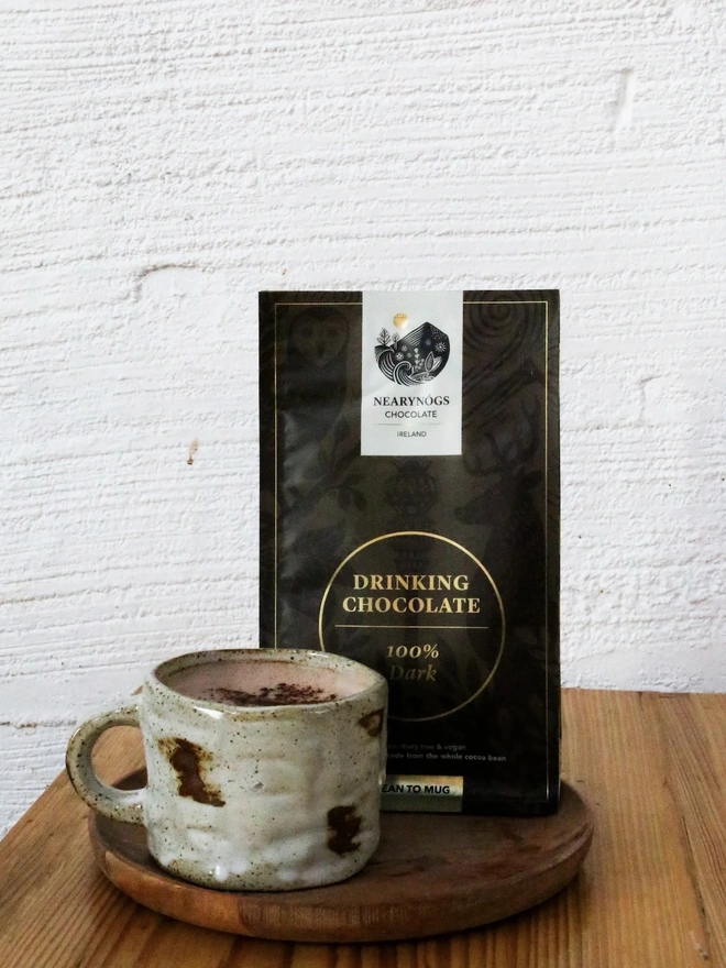 Drinking Chocolate 60% (Pack Of 200g/ 1kg), a hot chocolate sitting on a wooden tray on a kitchen table in front of a white backdrop. It is accompanied by a bag of drinking chocolate 