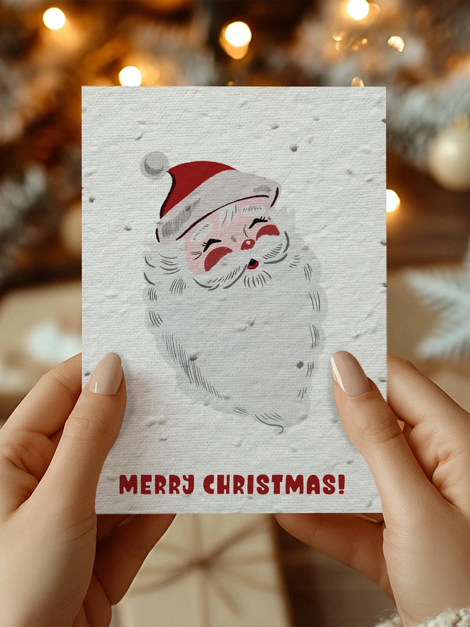 A pair of hands holding a Christmas card featuring an illustrated vintage Santa with 'Merry Christmas' underneath. The background has twinkly lights and presents.