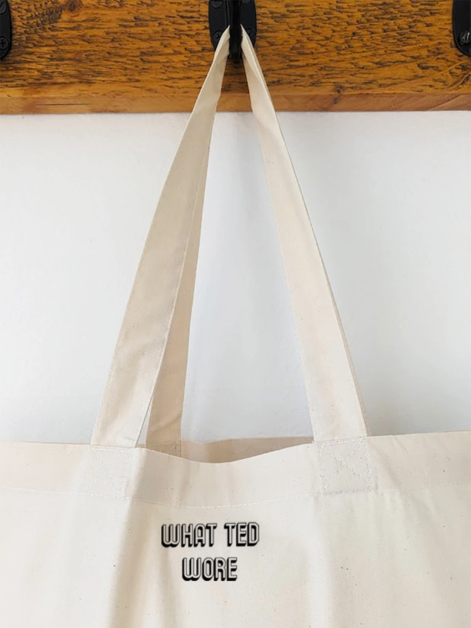 A hanging natural coloured tote bag with a black What Ted Wore logo