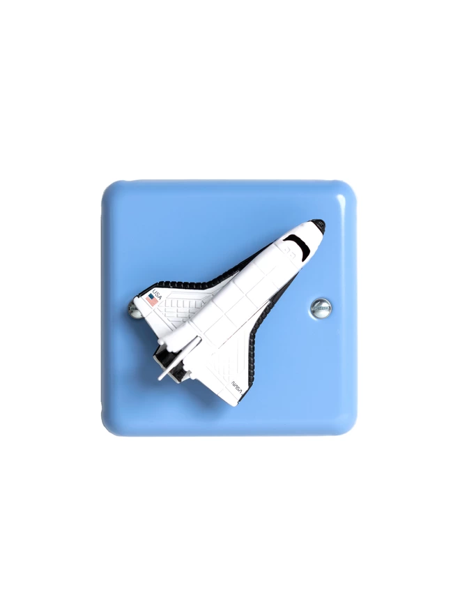 A pastel blue children's space themed dimmer light switch with a white space shuttle as the rotary knob to turn the lights on and off. The light switch plate is pale blue and made of metal, epoxy coated steel by Varilight. The space rocket is made of metal. The children's light switch brand is Candy Queen Designs.