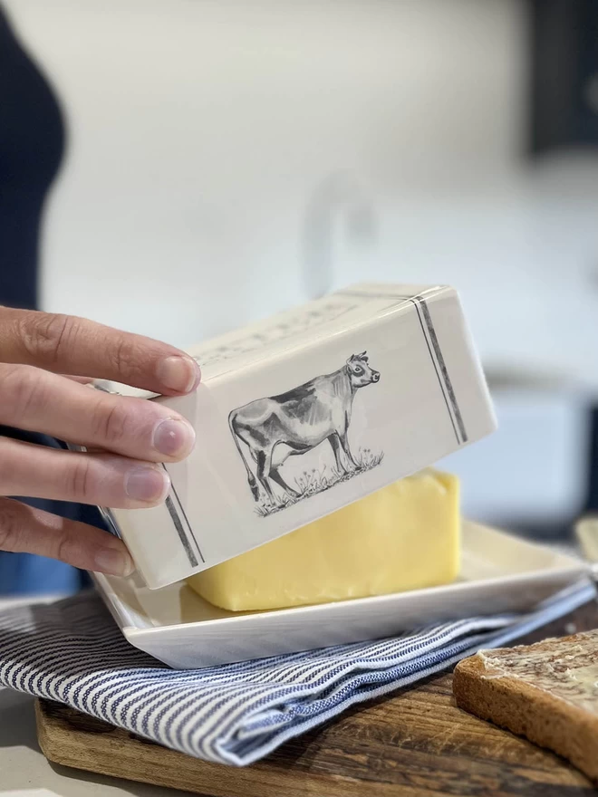  A handmade ceramic butter dish lid is being place back on its oblong plate over a block of butter.