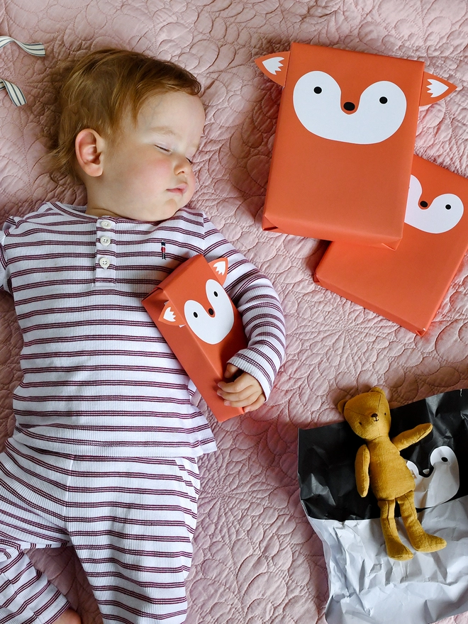 A baby wearing red and white striped pyjamas is asleep on a pink quilt with several gifts wrapped as foxes lay beside.