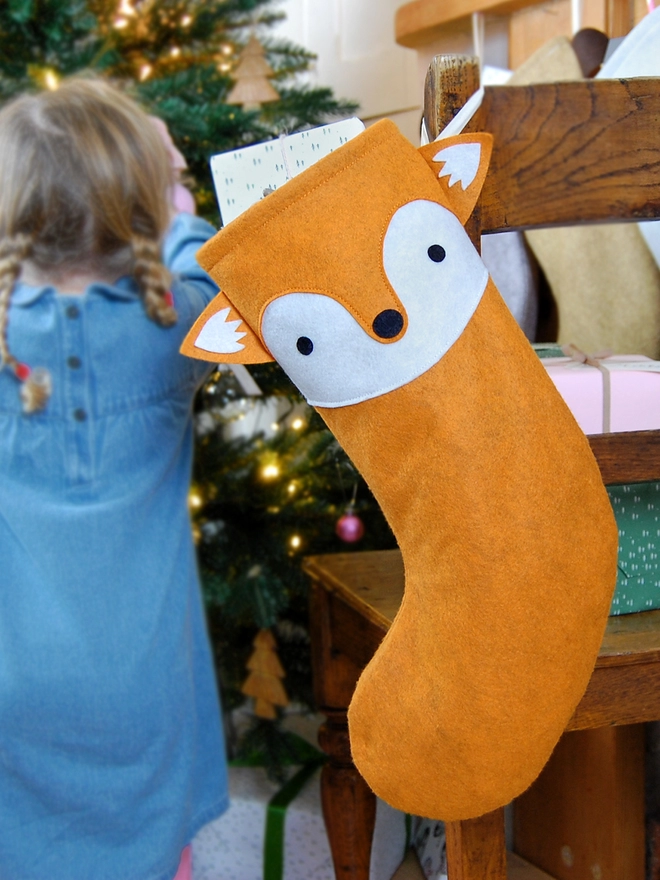 A handmade felt fox stocking hangs on a wooden chair, where a little girl sits, in front of a Christmas Tree.