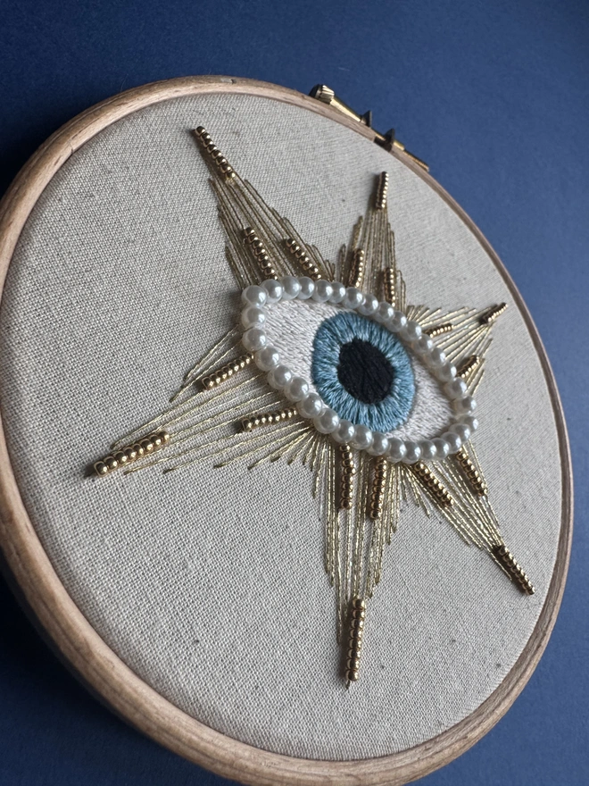 Embroidery hoop containing a blue eye edged with pearl beads and a gold star with gold beads
