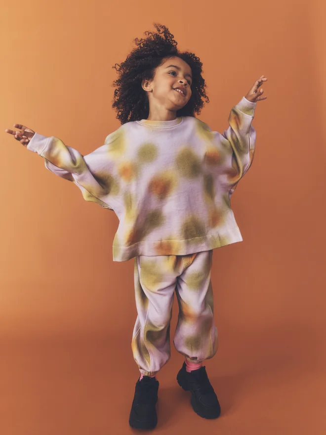 Child dances with arms out against a burnt orange backdrop, wearing the Studio Koter Riverson top in Hazy Lilac with matching print joggers.