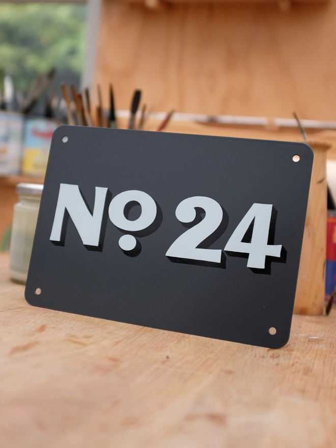 Hand painted house number in flourish style, pale blue and black No. 24 on anthracite grey metal plaque, in the artist's workshop