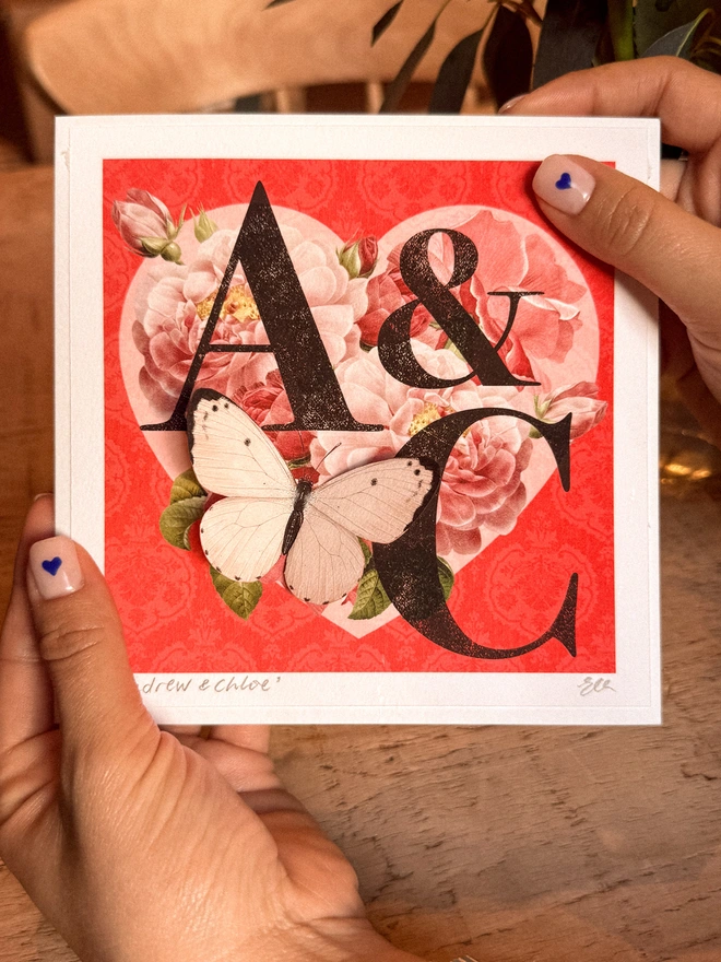 A handmade Ruby anniversary card featuring two initials of a couples names, place inside a heart design with vintage roses and on a red background. The recipients initials are hand written on the front of card and hand signed, A pink 3D paper cut butterfly sits on the heart and wings can be folded out for display.