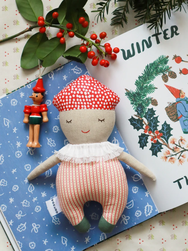 Soft handmade mushroom-themed doll with a red spotted cap, embroidered smiling face, ruffled white collar, red-striped body, and small green feet.
