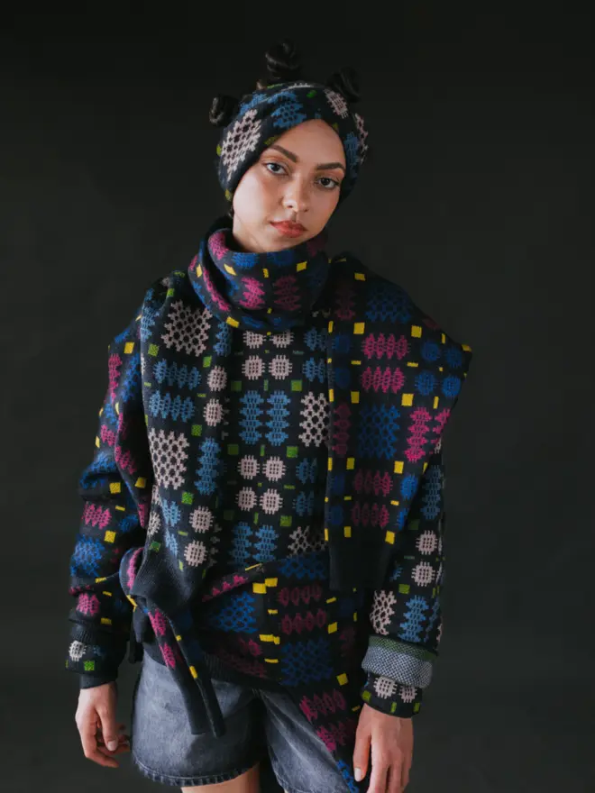 Model wearing pieces from MABLI's 'Carthen' collection, featuring the Carthen Wrap in the vibrant 'Emeralds' colour-way. The design showcases bold, colourful patterns inspired by traditional Welsh blanket tapestry.