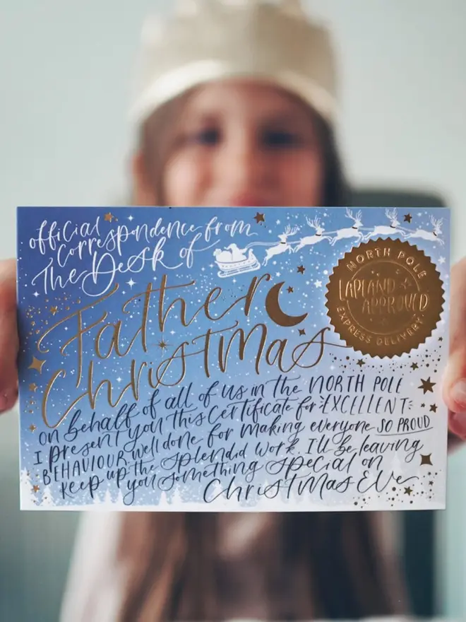 A child in a gold paper crown holding up the blue certificate with its gold seal.