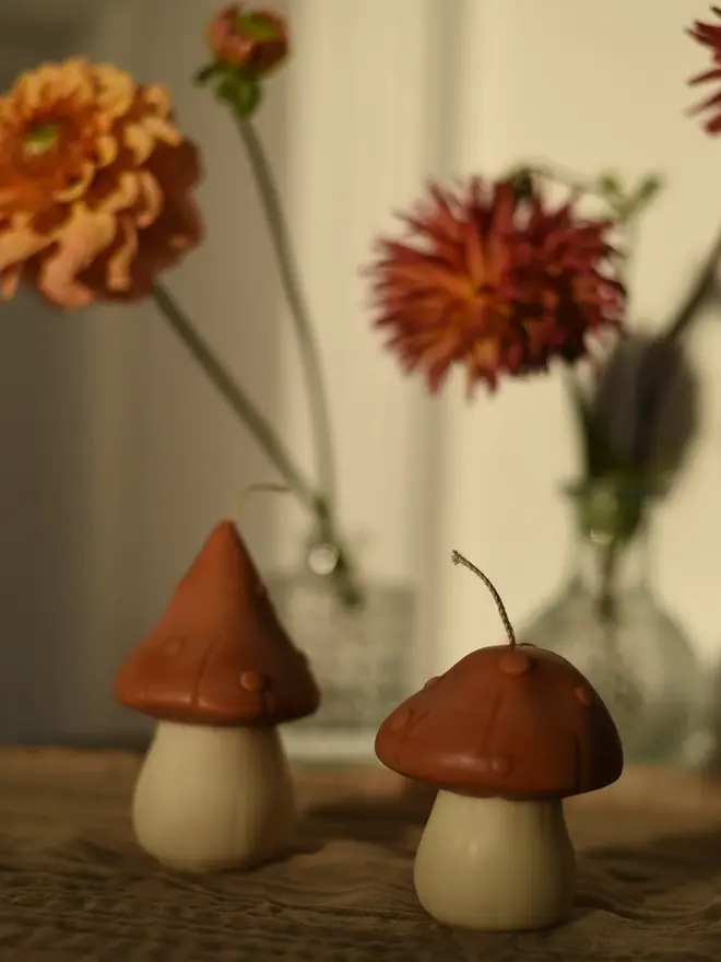 Pair Of Little Mushroom Candles (Set Of 2). A pair of terracotta mushroom candles with dotted caps and cream stems on a linen cloth, in front of orange and red dahlias in glass vases. The earthy, whimsical design brings woodland charm to autumn table styling.