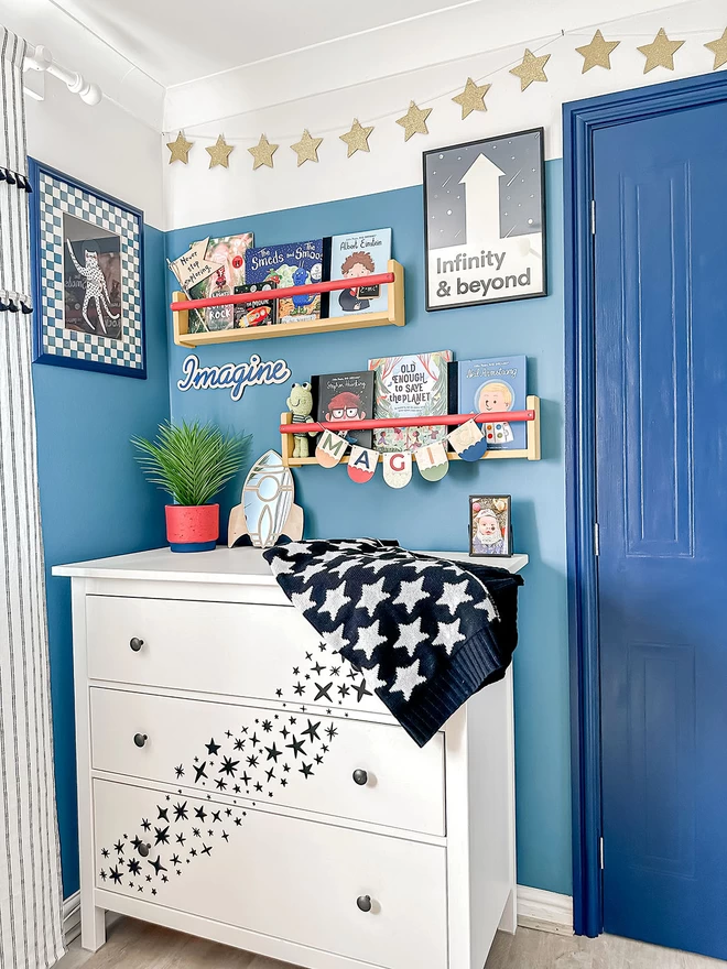 A view of a modern space themed nursery painted a vibrant blue. A white chest of drawers has a black and white star blanket draped over the side. Black star decals are stuck on to the front of the drawers cascading down. There are bookshelves above with childrens books and a print on the wall show a black and white typography print ‘to infinity and beyond’