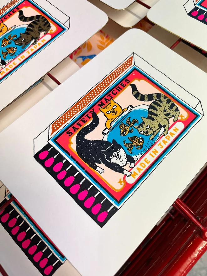 Three Cats And Two Fish Matchbox Lucky Dip, a group of colourful matchbox designs in the centre of white screenprints. They are grouped on a studio rack. 