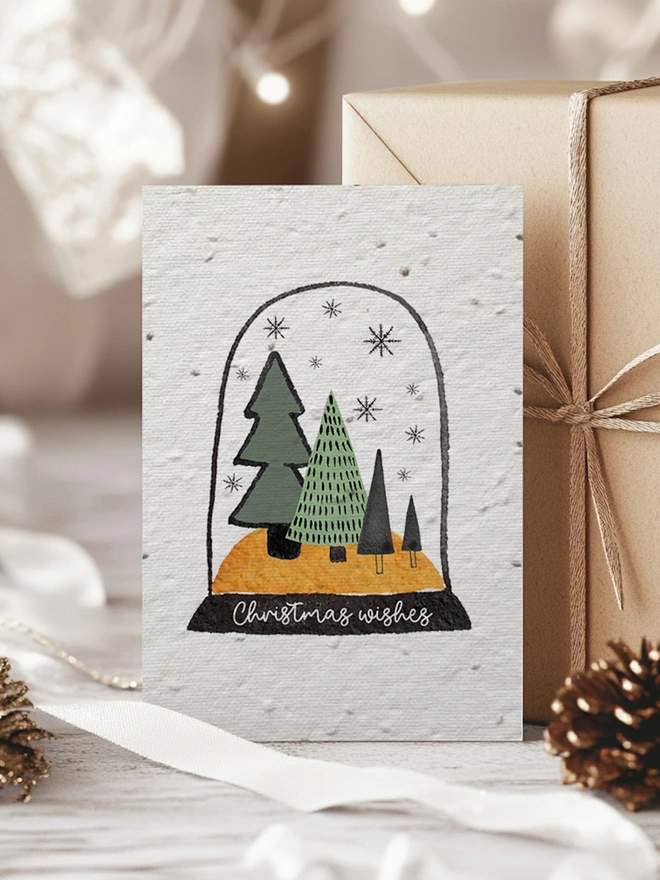 Christmas card with an illustrated snow globe on it with trees, snowflakes and 'Christmas Wishes'. The card is surrounded by presents, a pine cone and pine leaves.