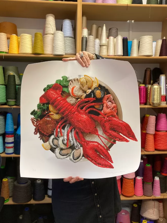 Kate holding the Fruits De Mer print in her studio with colourful threads seen stacked on shelves behind.