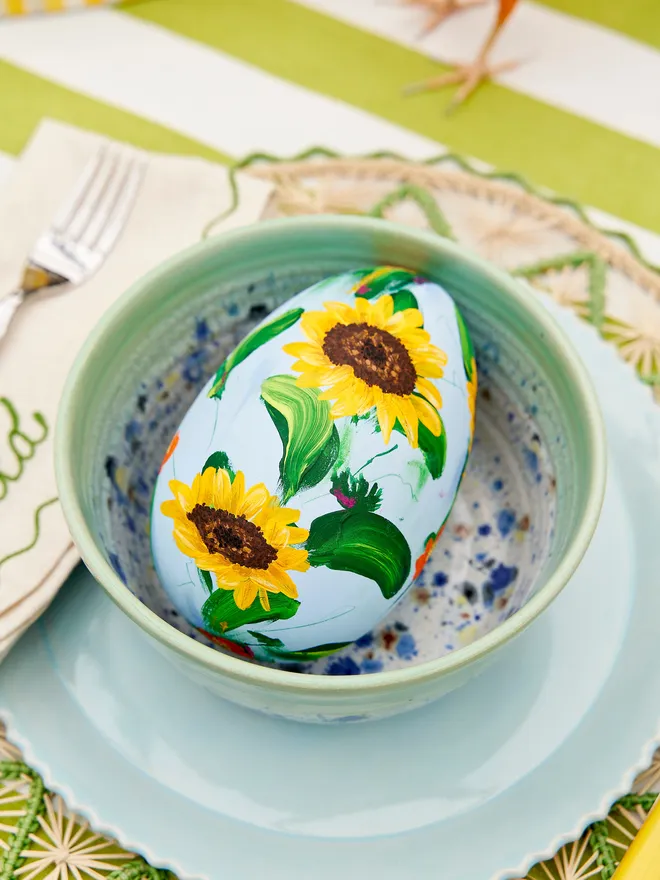 Soft blue hand painted wooden egg as part of a wider tablescape, positioned in a blue speckled bow on a blue scalloped plate. These are resting on a green jute tablemat atop a green and white striped tablecloth