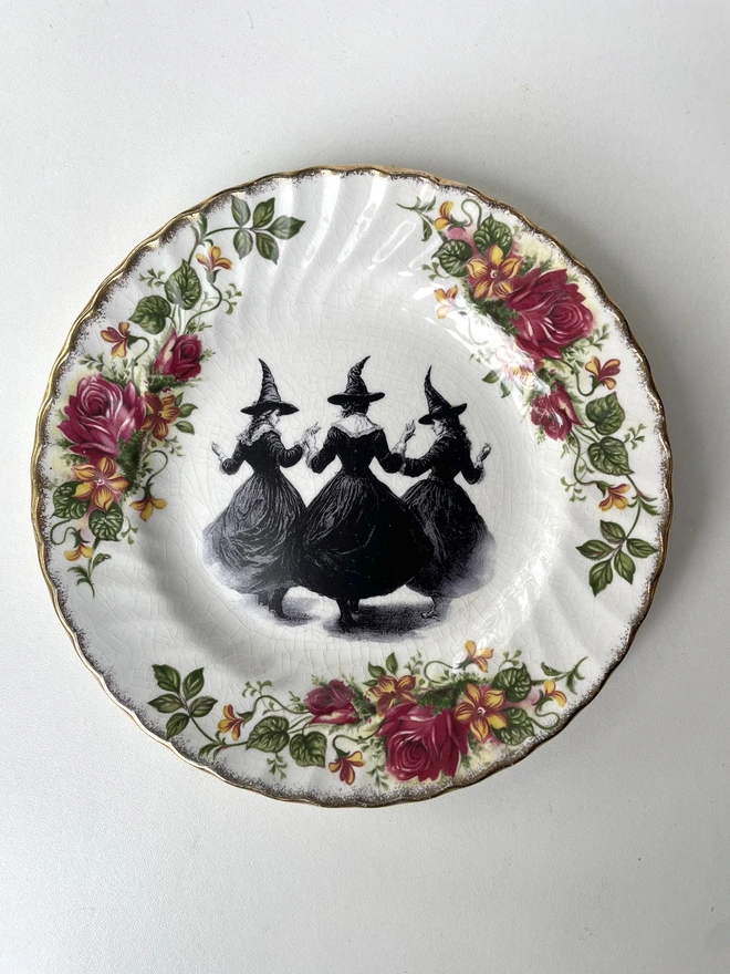 a coven of 3 witches dance hand in hand, the image is victorian in black and white and is printed directly onto an ornate vintage plate