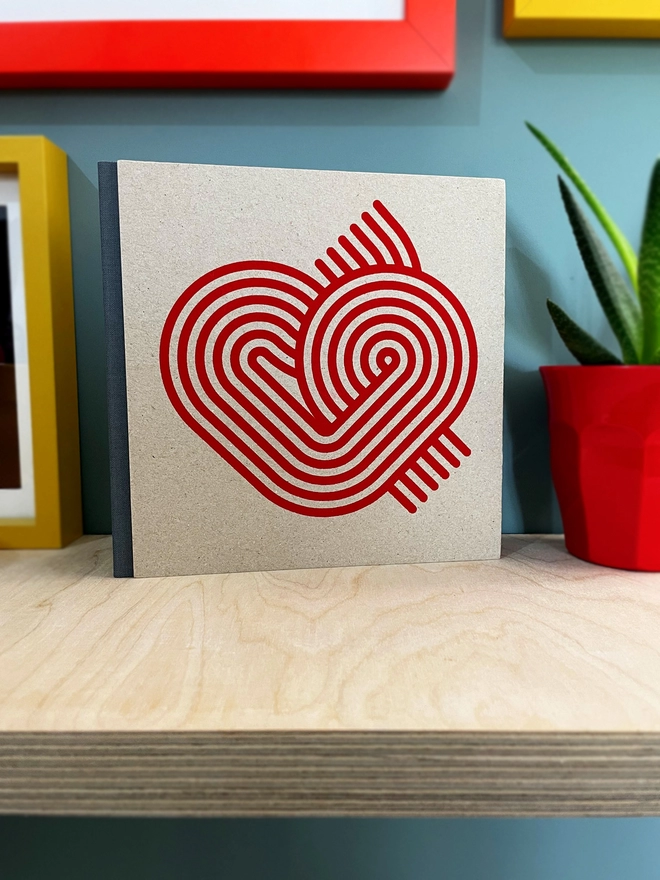 Stripy Heart design is screenprinted in magenta on a grey pasteboard sketchbook with a grey fabric spine, stood on a plywood shelf with hints of framed pictures around. the wall is duck egg blue, a plant glimpsed to the side.
