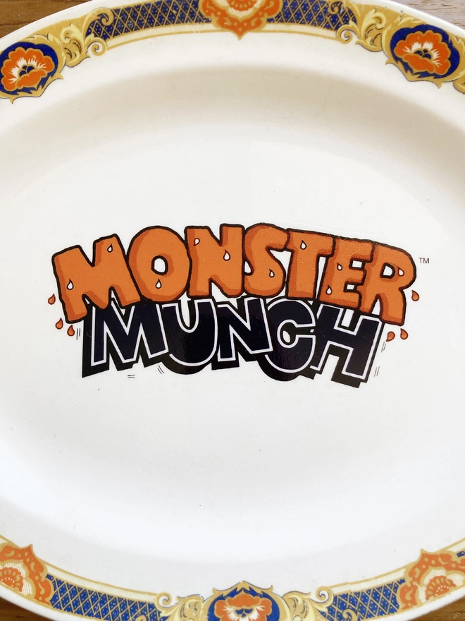 oval shaped vintage plate with the 'Monster Munch' logo in the centre. The rim of the plate is orange and dark blue, and the logo has been created to reflect these colours.