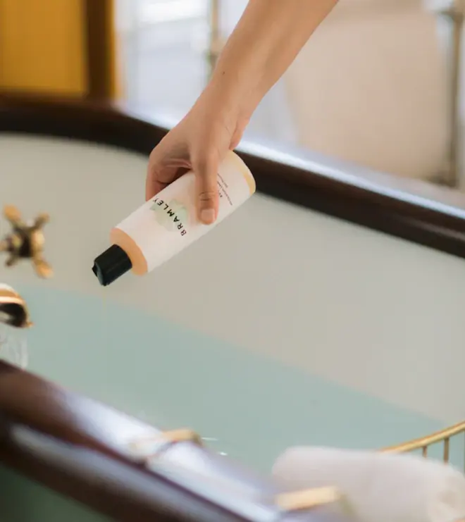 hand holding white bottle of a pink tinted bubble bath with black cap, pouring it into a bath tub full of water