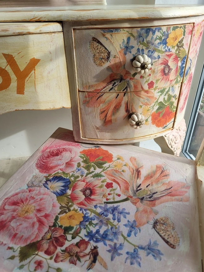 An antique kidney-shaped dressing table with chair and a trio of mirrors. Upcycled in cream and pale pink with decoupage florals in oranges and pinks. Text across the table read ‘flowers make me happy’ in the orange of the undercoat.