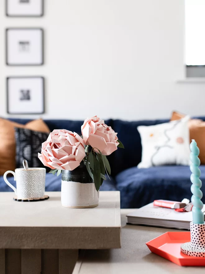 Two blush pink paper flowers in a vase on a coffee table, close to a sofa & other bits and pieces.