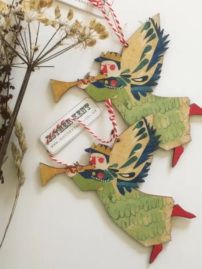 Joyful Angel illustrated wooden Christmas decoration in green, gold and red. Two decoratations lie on a white background alongside dried stalks.