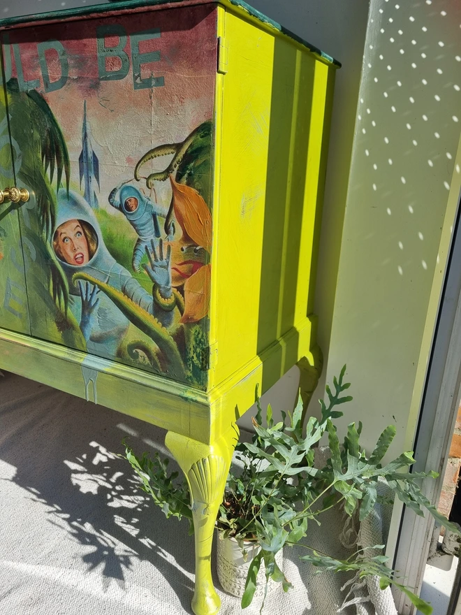 A painted record cabinet decorated with a sci fi image from the 1950s of a lady running from an alien and on the other door, a rose taken from a Victorian seed packet. There is wording that runs down the middle of the cabinet which reads 'it could be worse'. The colours are apple green and pinks.