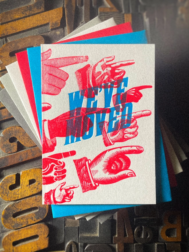 A double-sided, hand-printed letterpress artisan moving house announcement card with bold blue “We've Moved” typography and fluorescent red pointing hand illustrations (fists) on thick premium white card.